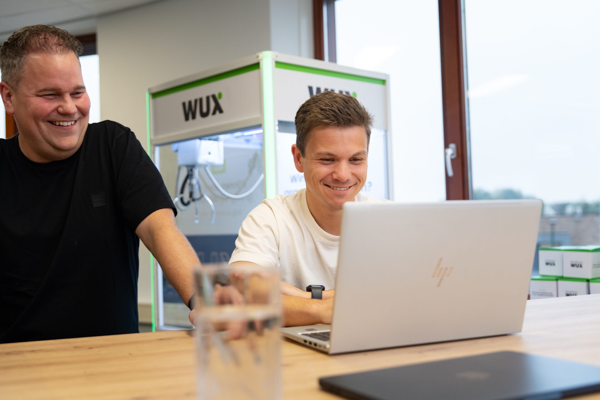 Twee specialisten van Wux AI lachen samen achter een laptop, symbool voor de succesvolle adoptie en training van medewerkers bij de implementatie van AI.